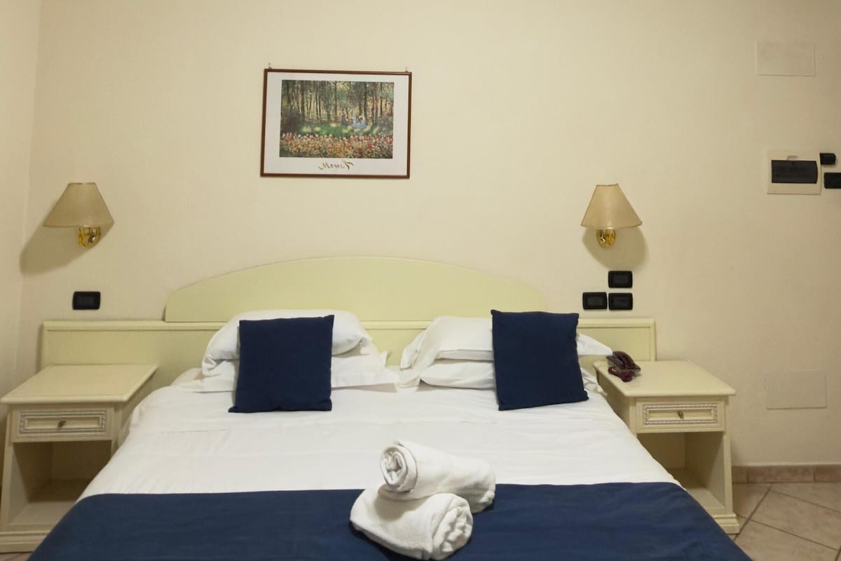 A hotel room with a bed, two bedside tables, a nightstand, a painting on the wall, and an air conditioning unit.