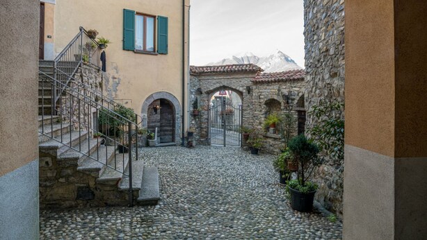 A cobblestone courtyard with stone buildings, a staircase, and a view of mountains. - GuestHost - Comfortable mezzanine apartment of approximately 35 square meters, accommodating up to four people, located on the first floor of a building without an elevator.The property is located in Visgnola, a quiet hamlet of Bellagio, one of Lake Como (Bellagio)