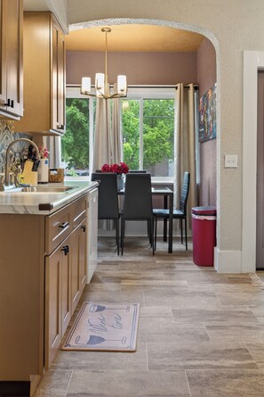 A kitchen with a marble countertop, wooden cabinets, a dining area with chairs, a red trash bin, and a view of greenery outside.
