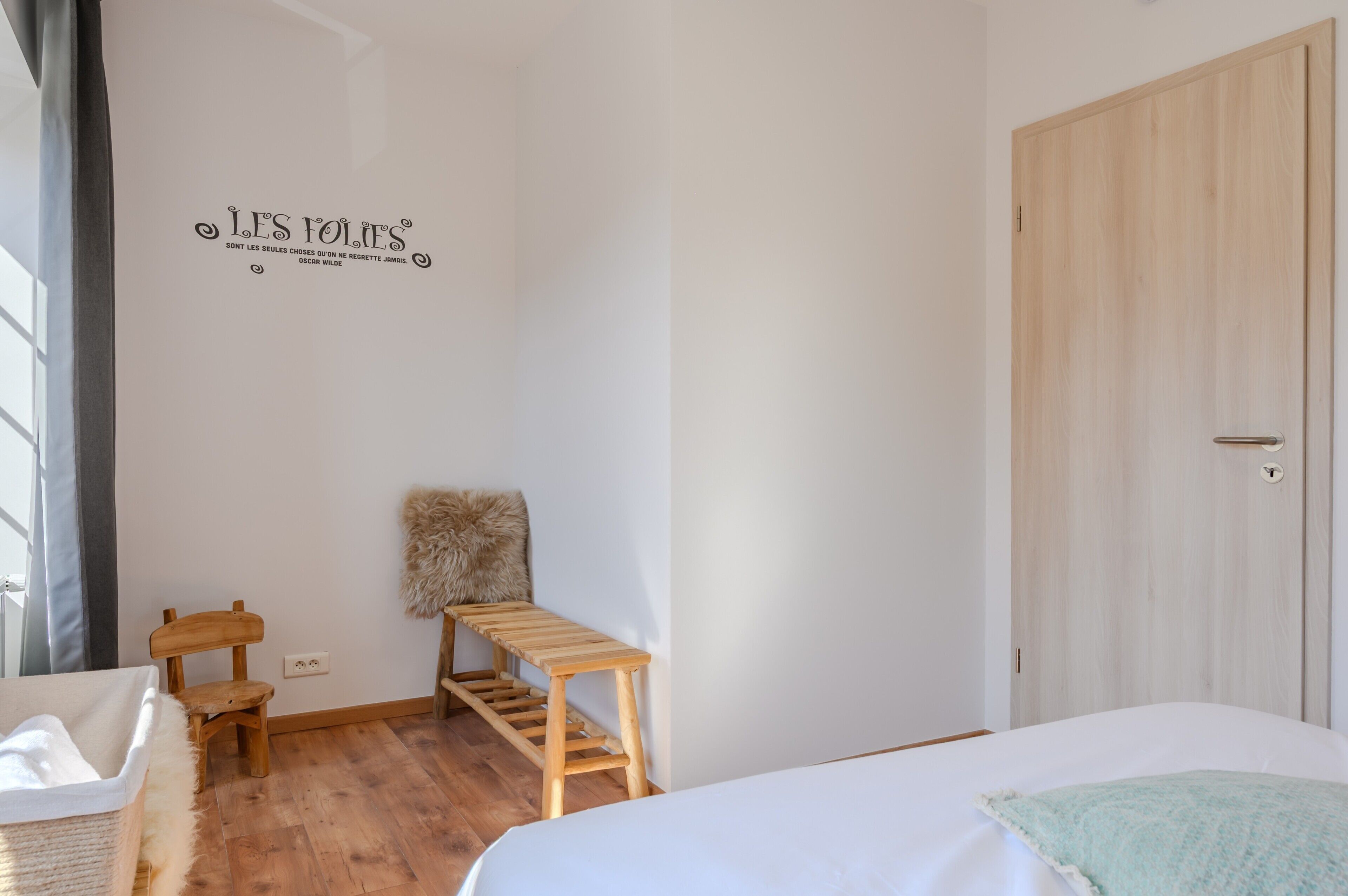 Une chambre avec un banc en bois, un plaid en fourrure, un lit blanc et une porte en bois.