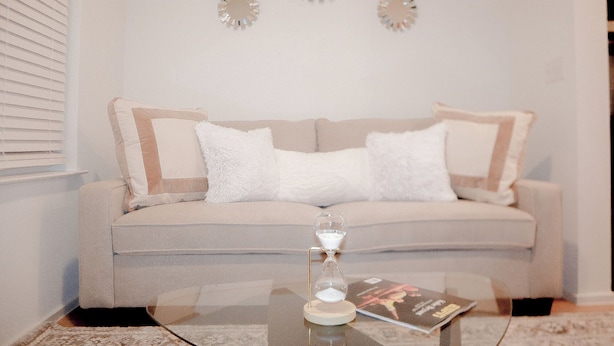 Een woonkamer met een beige bank, witte kussens en een glazen salontafel met een boek en een klein zandloper.