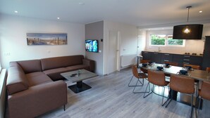 Un salon moderne avec un canapé marron, une table basse et un téléviseur. On y trouve également un coin repas avec une longue table et des chaises. Les murs sont blancs et le sol est en bois.
