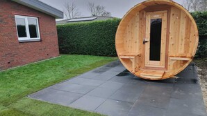Une structure de sauna en bois, dotée d’une porte, est installée sur une surface pavée, à côté d’un bâtiment en briques et d’une haie.