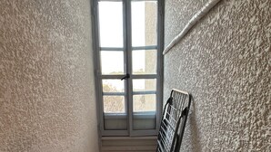 Une pièce étroite avec un parquet en bois disposé en chevrons, une fenêtre aux cadres blancs et un étagère en métal adossée à un mur à la texture particulière.