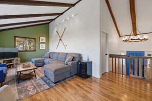 Uma sala de estar moderna com sofá seccional, piso de madeira e teto com vigas aparentes.