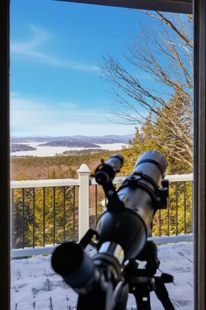 Um telescópio numa varanda com vista para uma paisagem nevada com árvores e um corpo de água.
