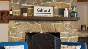 A stone fireplace with a sign reading "Gilford" and coordinates. Two blue armchairs with checkered pillows are in front. - Lake Views & Mountain Adventures Await (Gilford)