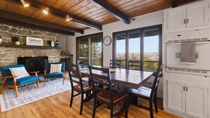 A cozy dining area with a stone fireplace, wooden ceiling beams, and a view through large windows. - Lake Views & Mountain Adventures Await (Gilford)