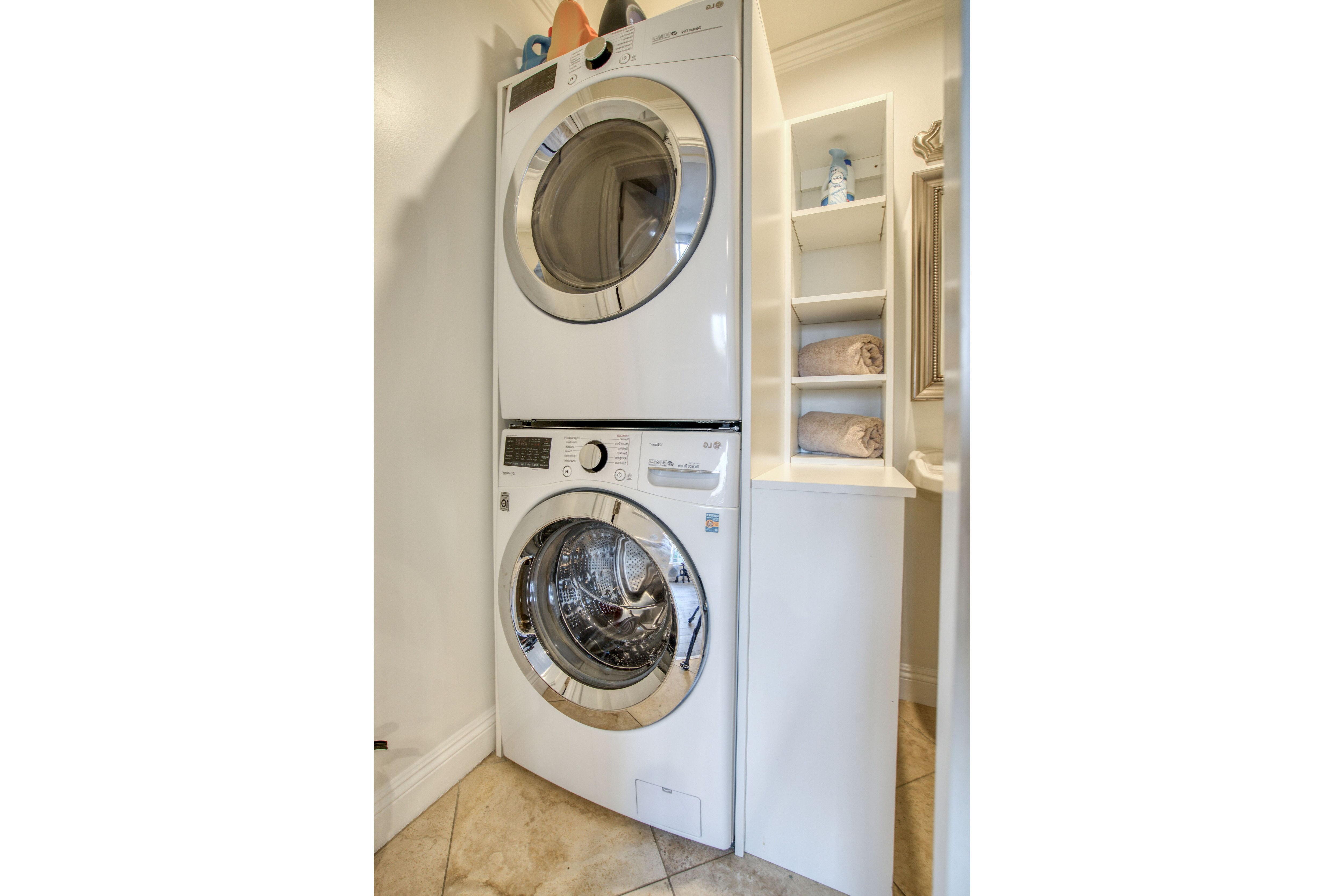 A front-loading washer and dryer unit in a laundry room with a tiled floor and a white shelving unit to the right.
