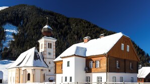 A traditional wooden building with a snow-covered roof, adjacent to a church with a bell tower, set against a backdrop of a snow-covered mountain and clear blue sky. - Heavenly vacation - living in the vicarage (Sölk)