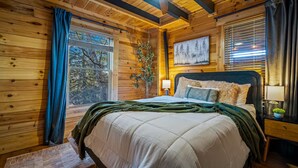 Ein gemütliches Schlafzimmer in einer Holzhütte mit einem großen Bett, einem Fenster mit Vorhängen, einem Bild an der Wand und einem Nachttisch mit Lampe.