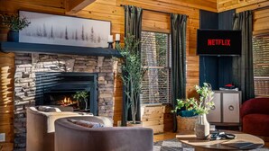 Ein gemütliches Blockhauss-Interieur mit Kamin aus Stein, bequemen Sitzmöglichkeiten, einem Fernseher und einem großen Fenster mit Vorhängen.