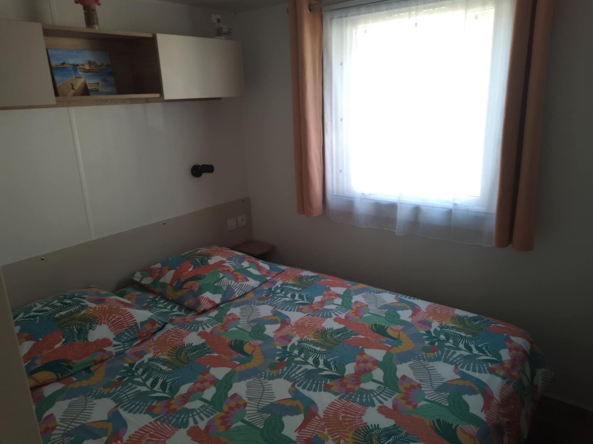 A bedroom with a bed, a window with curtains, a shelf with a picture, and a door.