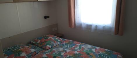A bedroom with a bed, a window with curtains, a shelf with a picture, and a door.