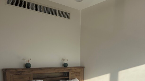 Una cama bien hecha con sábanas blancas y cabecera de madera, un florero con flores secas sobre la cabecera y un respiradero en la pared.