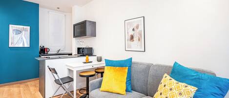 A compact living space with a kitchenette, a grey sofa with colorful cushions, a small dining table, and a foldable chair.