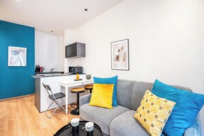 A compact living space with a kitchenette, a grey sofa with colorful cushions, a small dining table, and a foldable chair. - Guesthost - Dimora Ponte Vecchio Lovely Apartment (Florence)