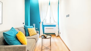Un salon moderne avec un canapé gris, des coussins turquoise et jaunes, une petite table d’appoint noire et une fenêtre ornée de rideaux blancs.