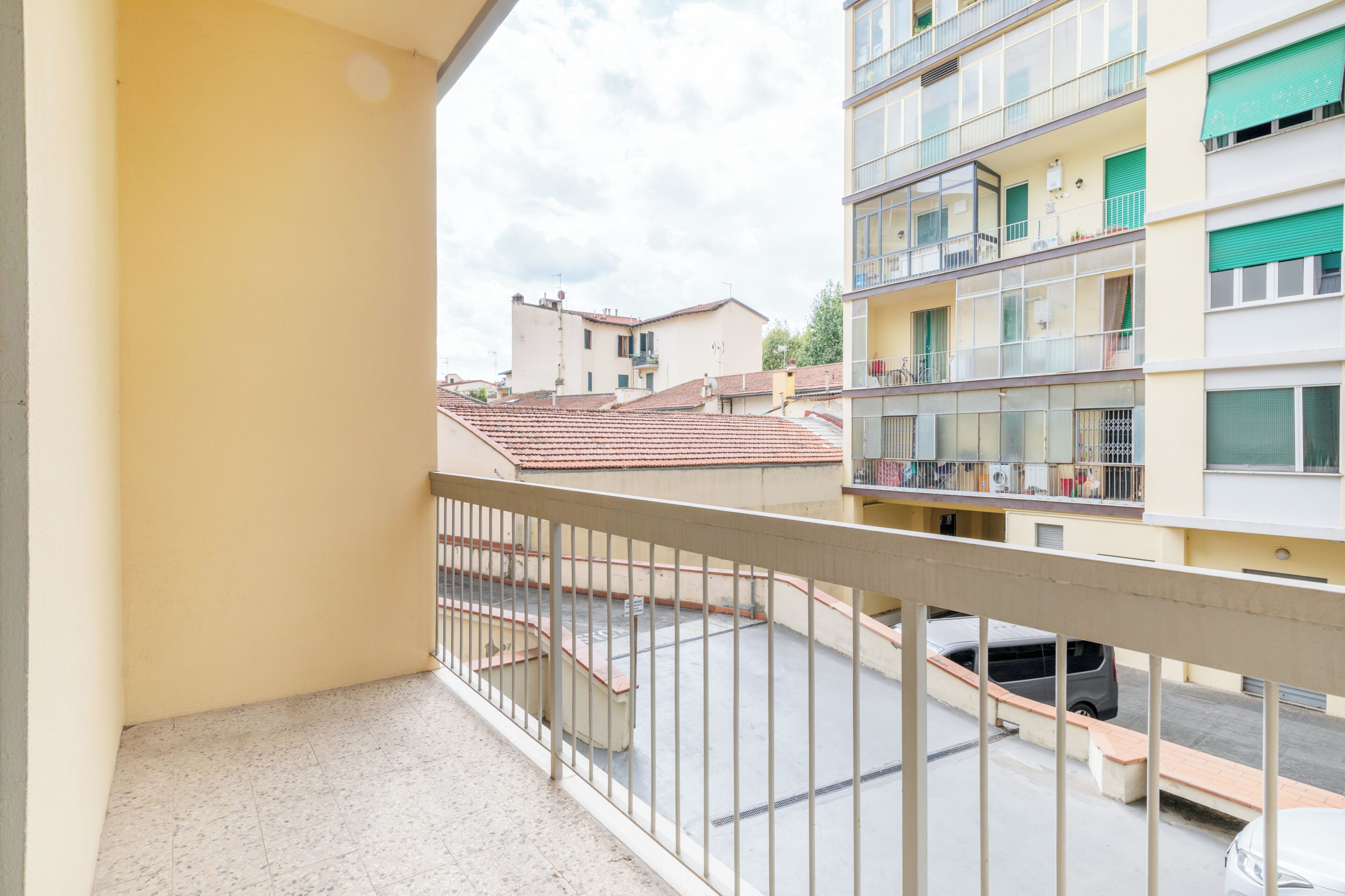 A balcony with a metal railing, a view of a residential area with multi-story buildings, and a clear sky.