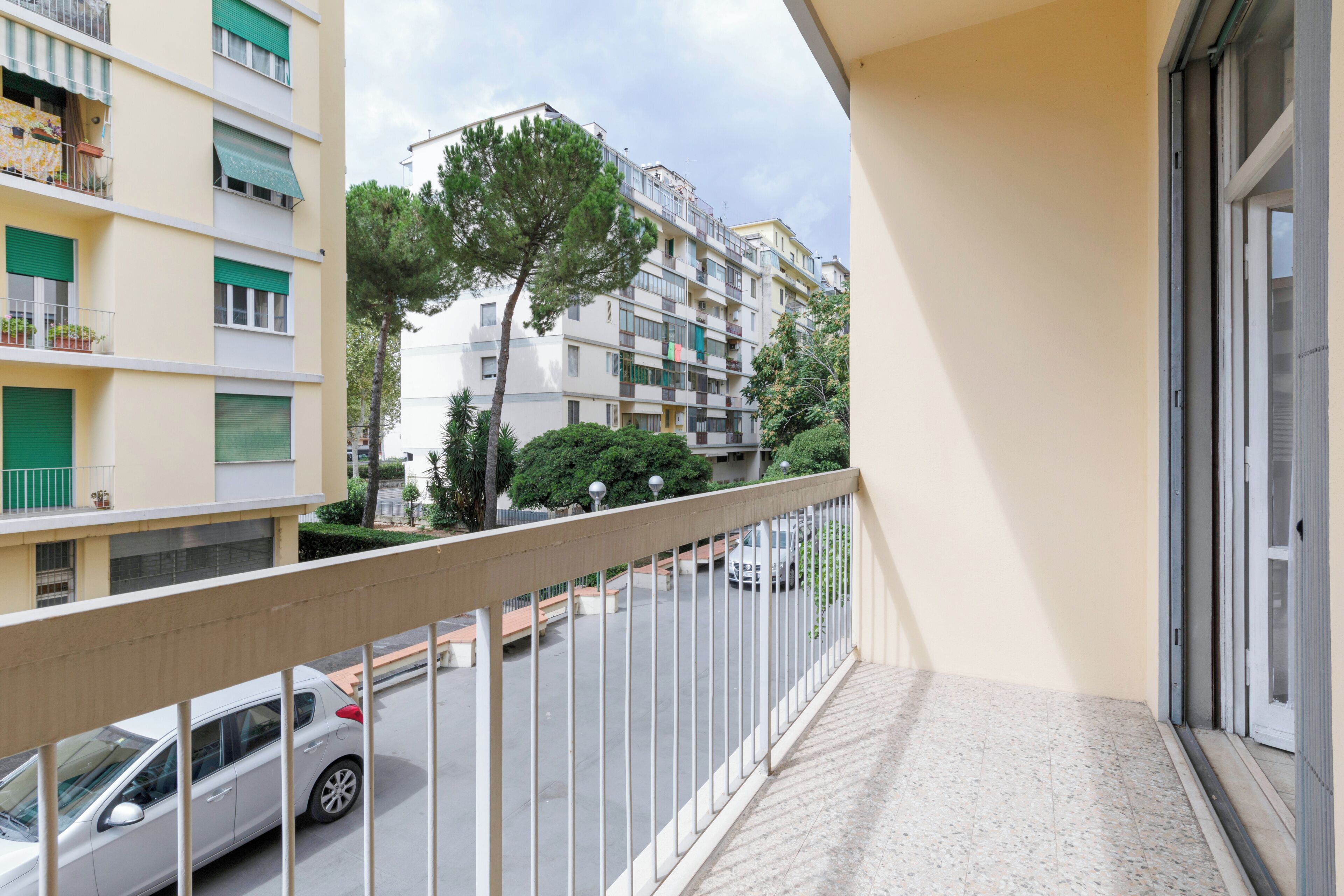 A balcony with a metal railing overlooking a street with parked cars and a multi-story building with balconies.