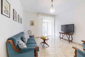 A living room with a sofa, a television, a table with flowers, and framed pictures on the wall. - Guesthost - Il Salotto Blu - Appartamento Con Parcheggio (Florence)