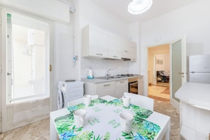 A compact kitchen with a washing machine, a dining table with four white mugs, and a view into the living area. - Guesthost - Il Salotto Blu - Appartamento Con Parcheggio (Florence)