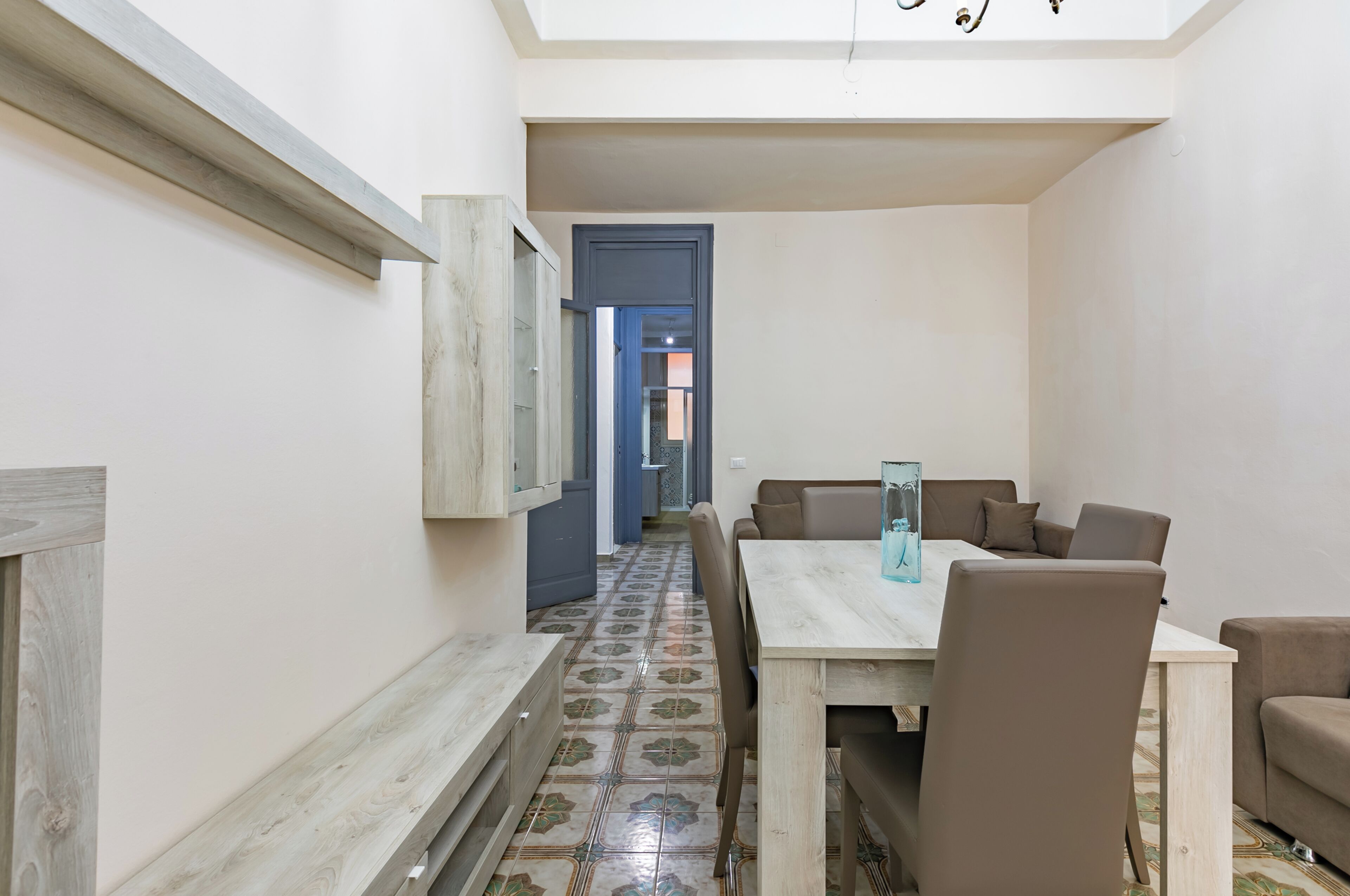 A narrow hallway leading to a dining area with a tiled floor, a wooden table, and brown chairs.