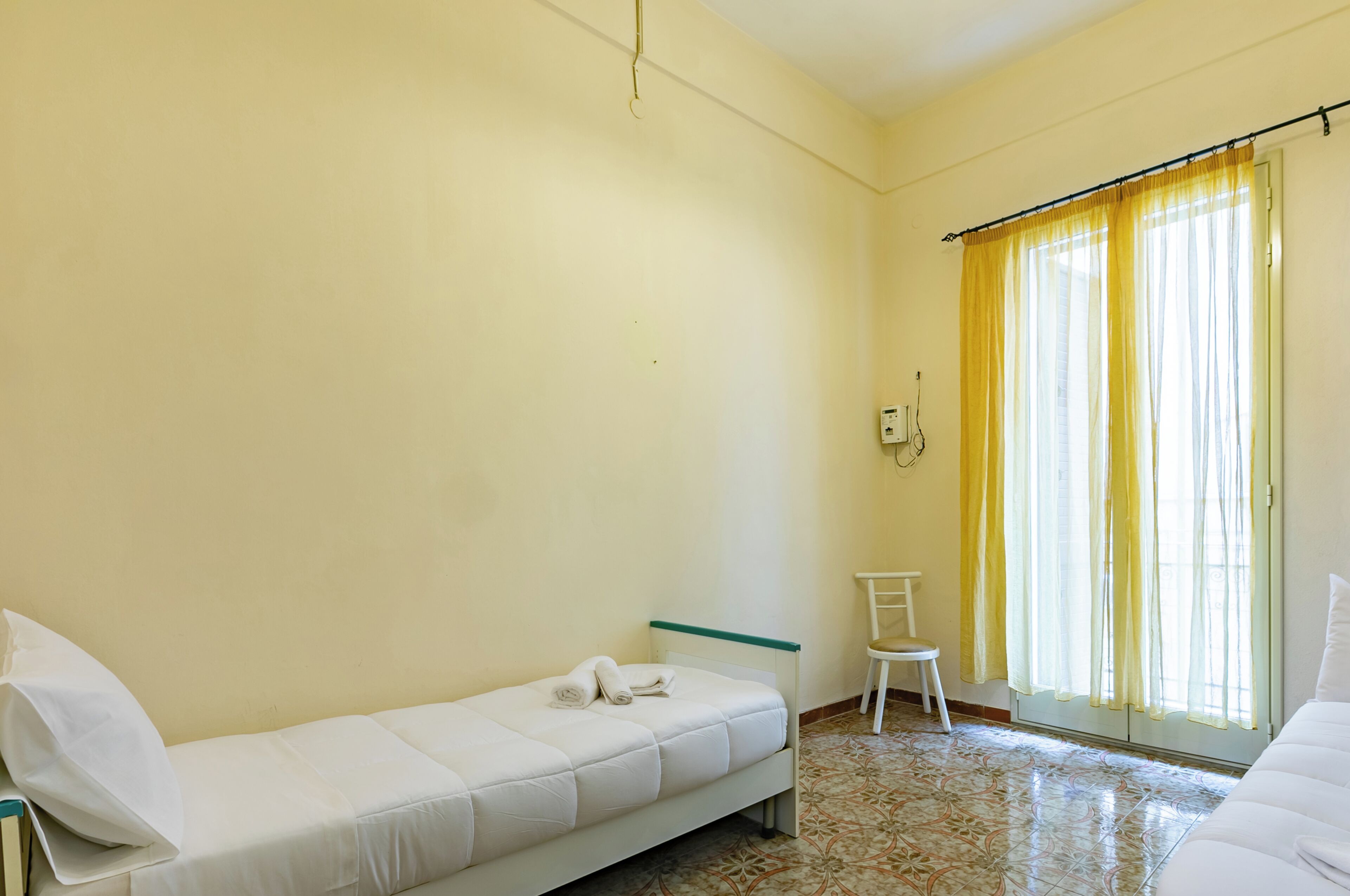 Une chambre avec un lit simple, une chaise, une table de chevet et une fenêtre avec des rideaux jaunes.