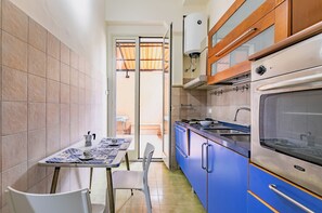 A compact kitchen with a dining area, featuring a table with a blue and white patterned tablecloth, a chair, a stove, and cabinets. - Guesthost - Teatro Massimo Roomy Flat With Covered Terrace (Palermo)