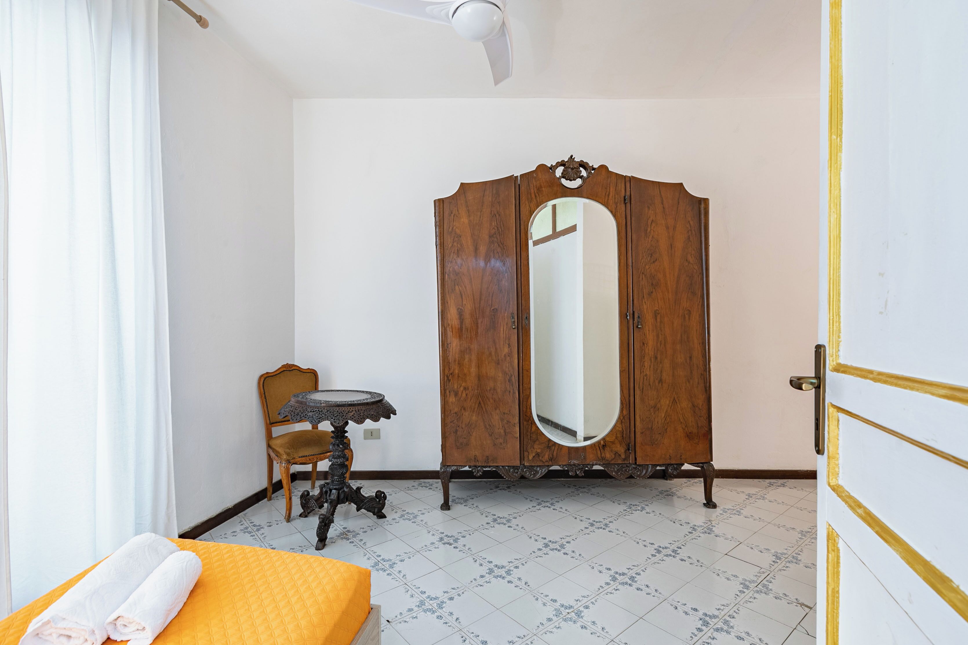 A bedroom with a bed, a wooden wardrobe with a mirror, a small round table, and a chair.