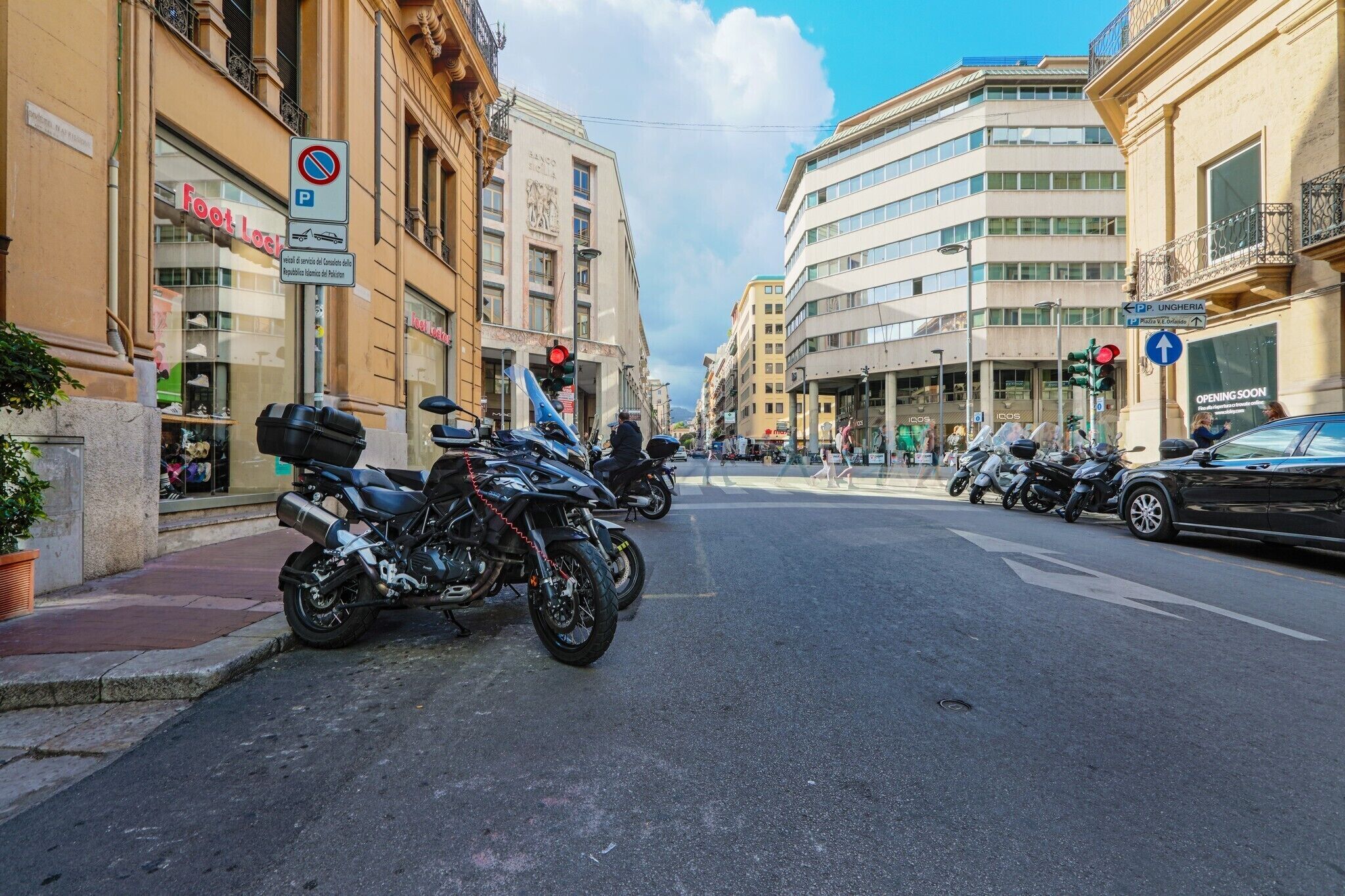 A street with parked motorcycles, a 'Foot Locker' store, and a building with 'OPENING SOON' signage.