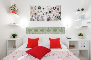 A modern bedroom with a bed, red pillows, a decorative wall piece, and white shelving with plants. - Guesthost - Fortezza Functional Studio (Florence)