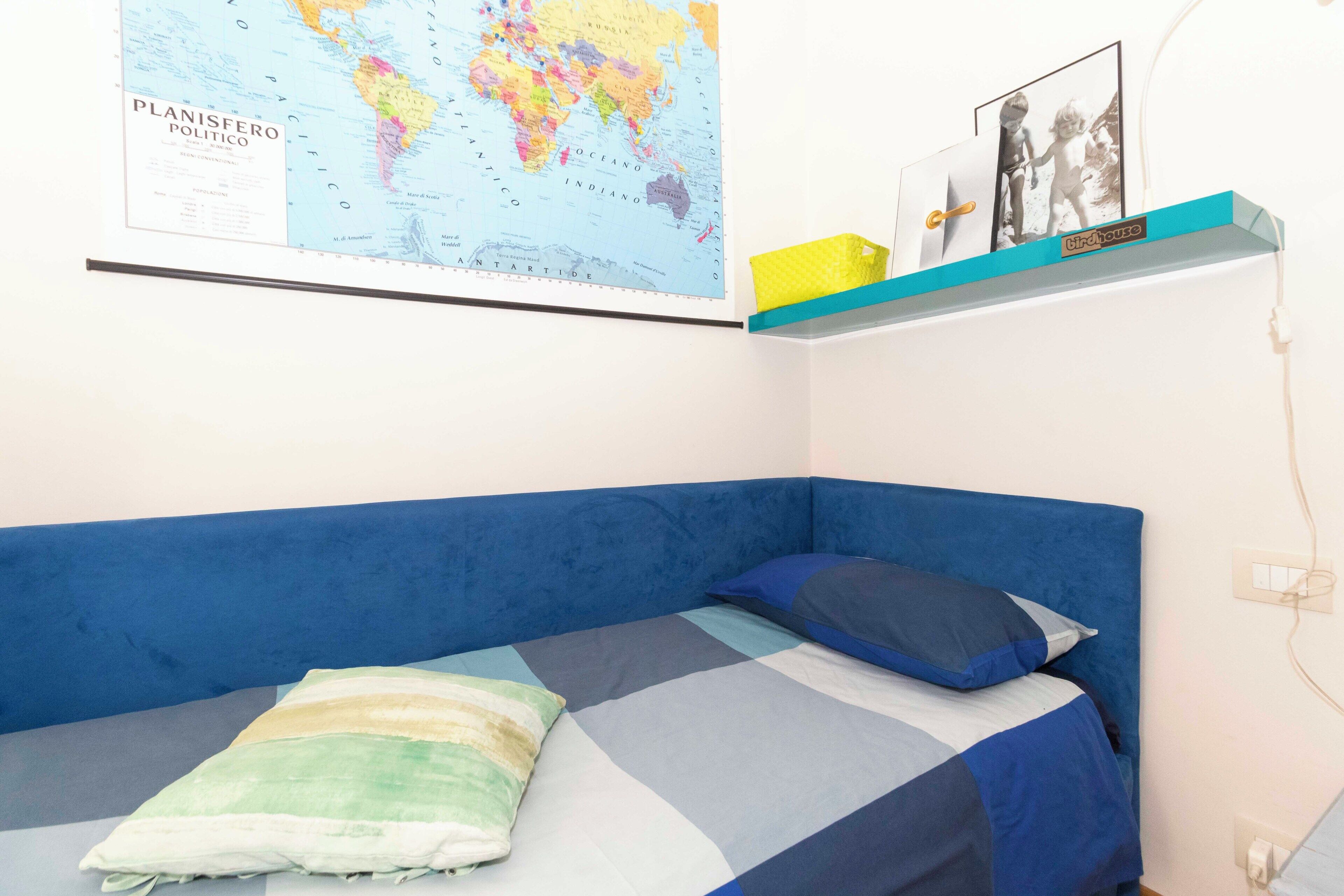 A room with a blue bed, a world map on the wall, and a shelf with framed photos.