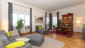Ein Wohnzimmer mit grauem Sofa, Hocker, Esstisch mit Stühlen, einem Holzschrank und einem Blumentopf.