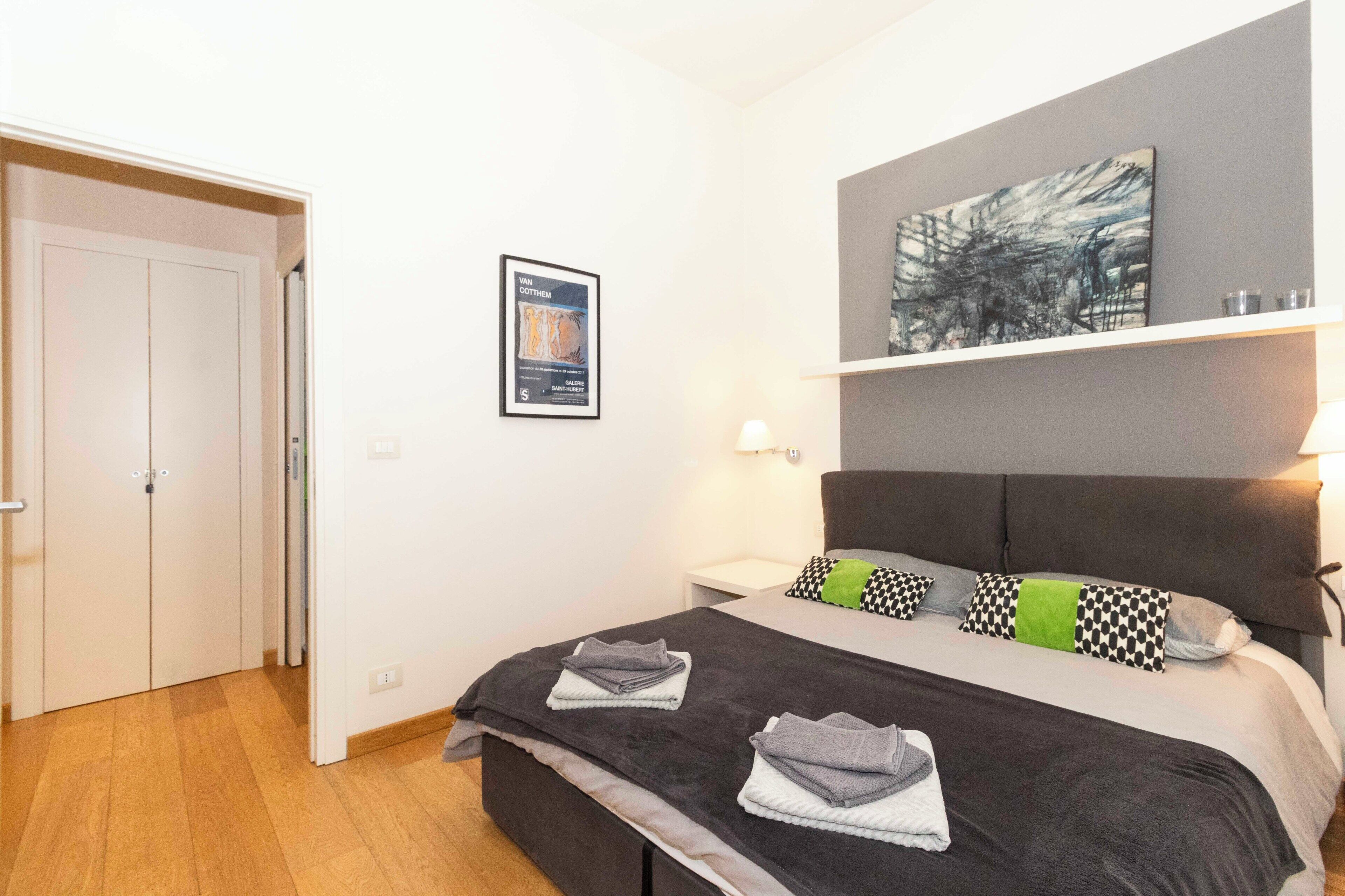 A bedroom with a bed, a nightstand, a wall-mounted shelf, a framed picture, and a door.