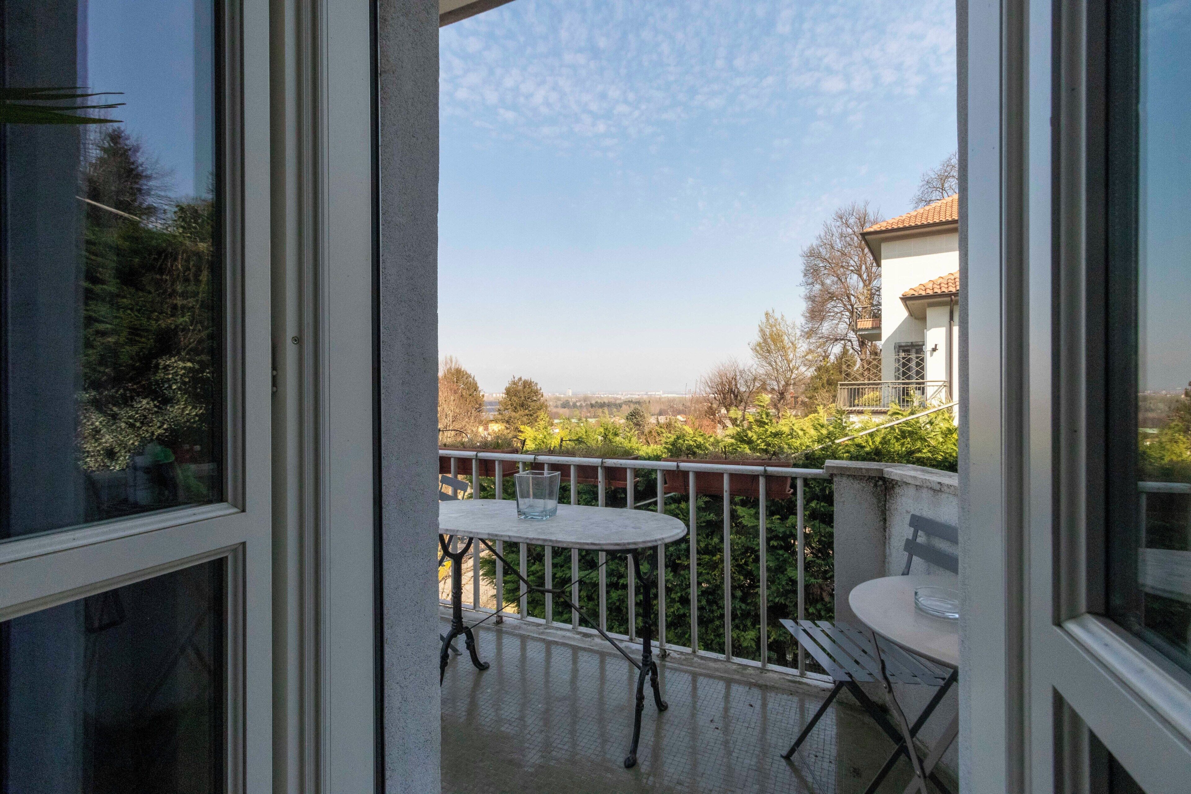 A balcony with a table and chairs, a glass of water, and a view of a building and trees.