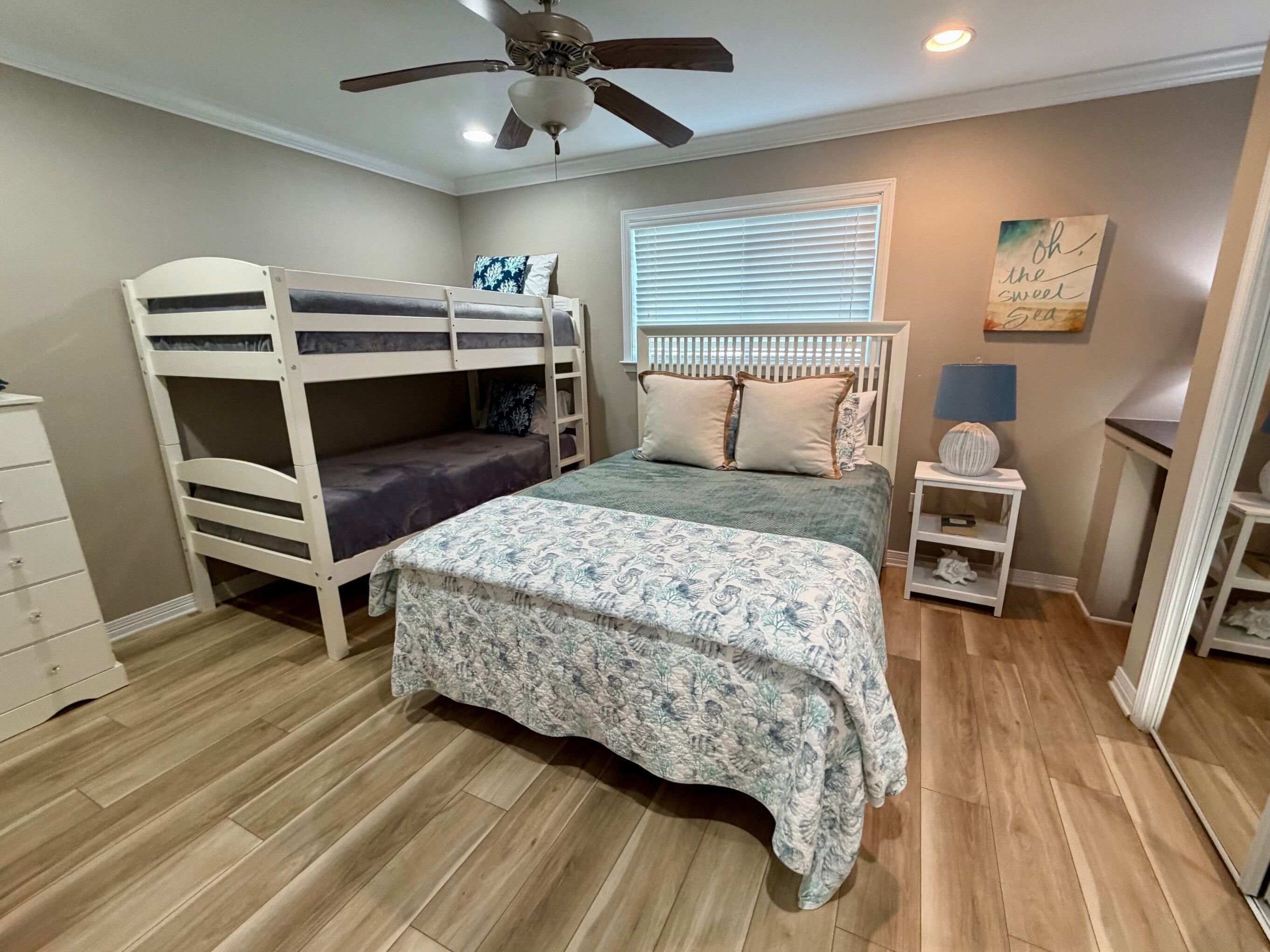 A bedroom with bunk beds, a ceiling fan, a window with blinds, a bedside table, and a dresser.