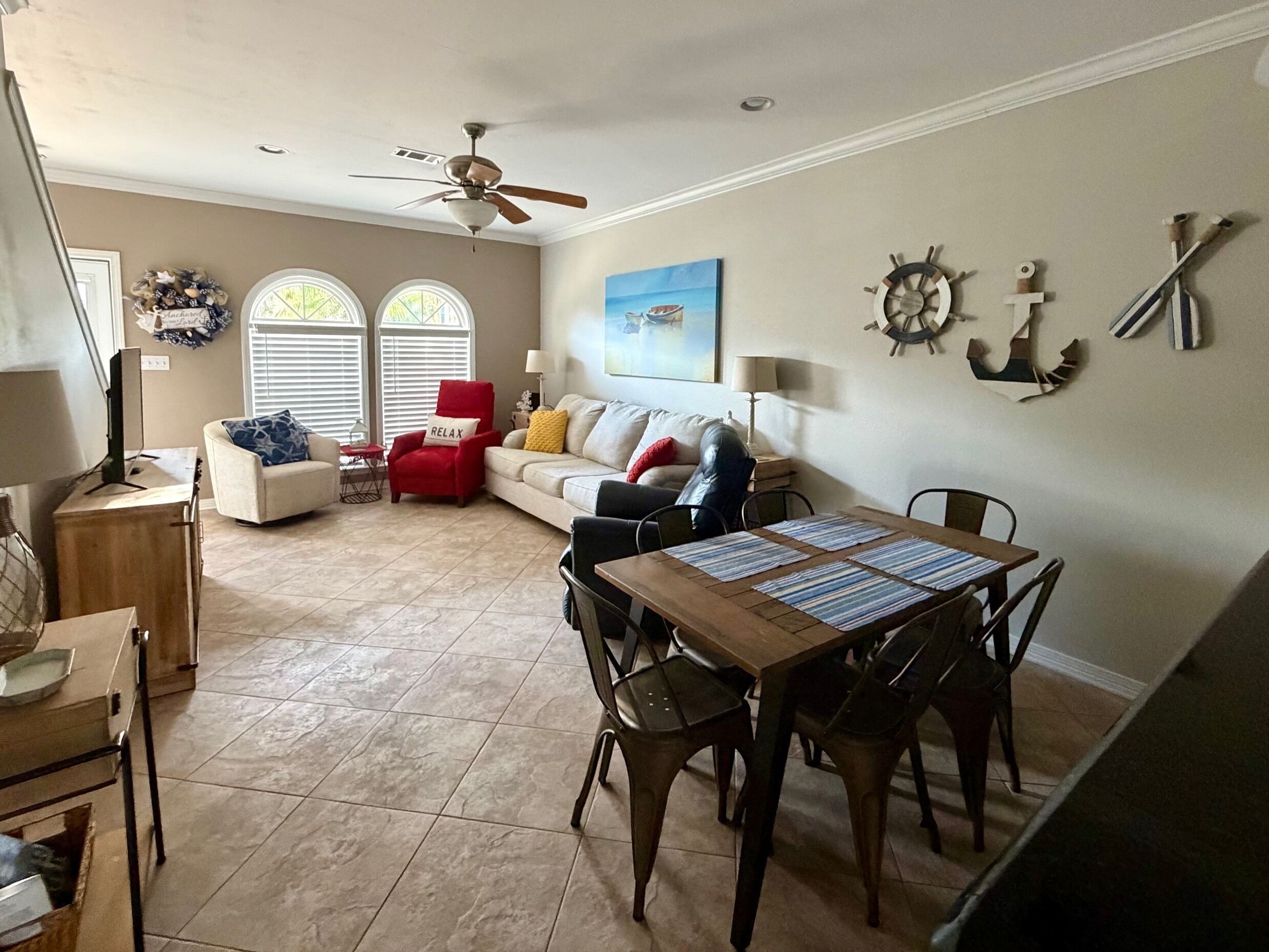 A living room with a dining area, a ceiling fan, a window with blinds, and a wall-mounted ship's wheel decoration.