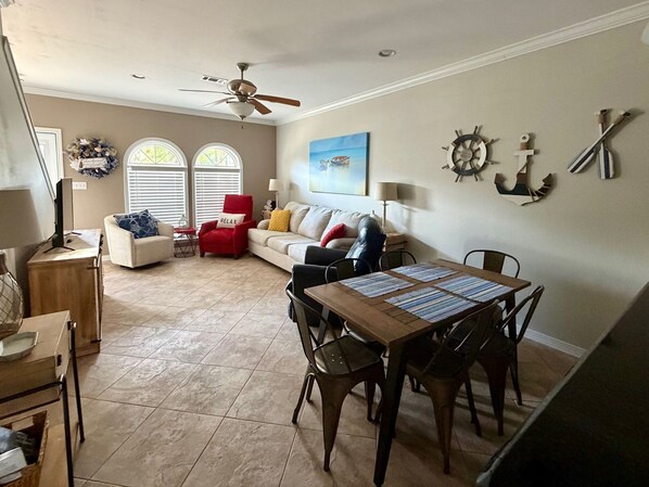 A living room with a dining area, a ceiling fan, a window with blinds, and a wall-mounted ship's wheel decoration.