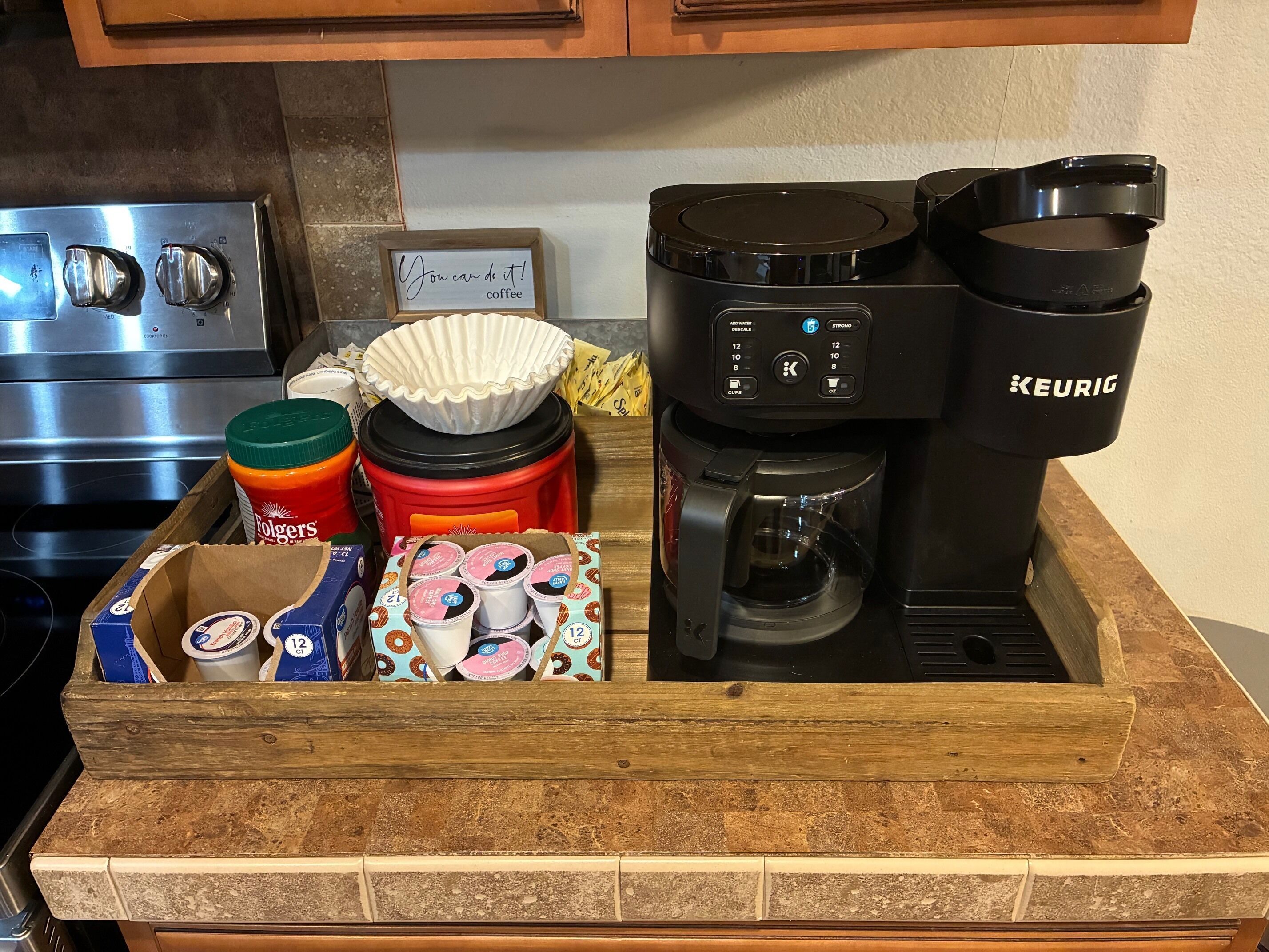 A kitchen counter with a coffee maker, a box of Folgers coffee, a jar of Folgers coffee, a coffee pod holder, and a coffee cup.