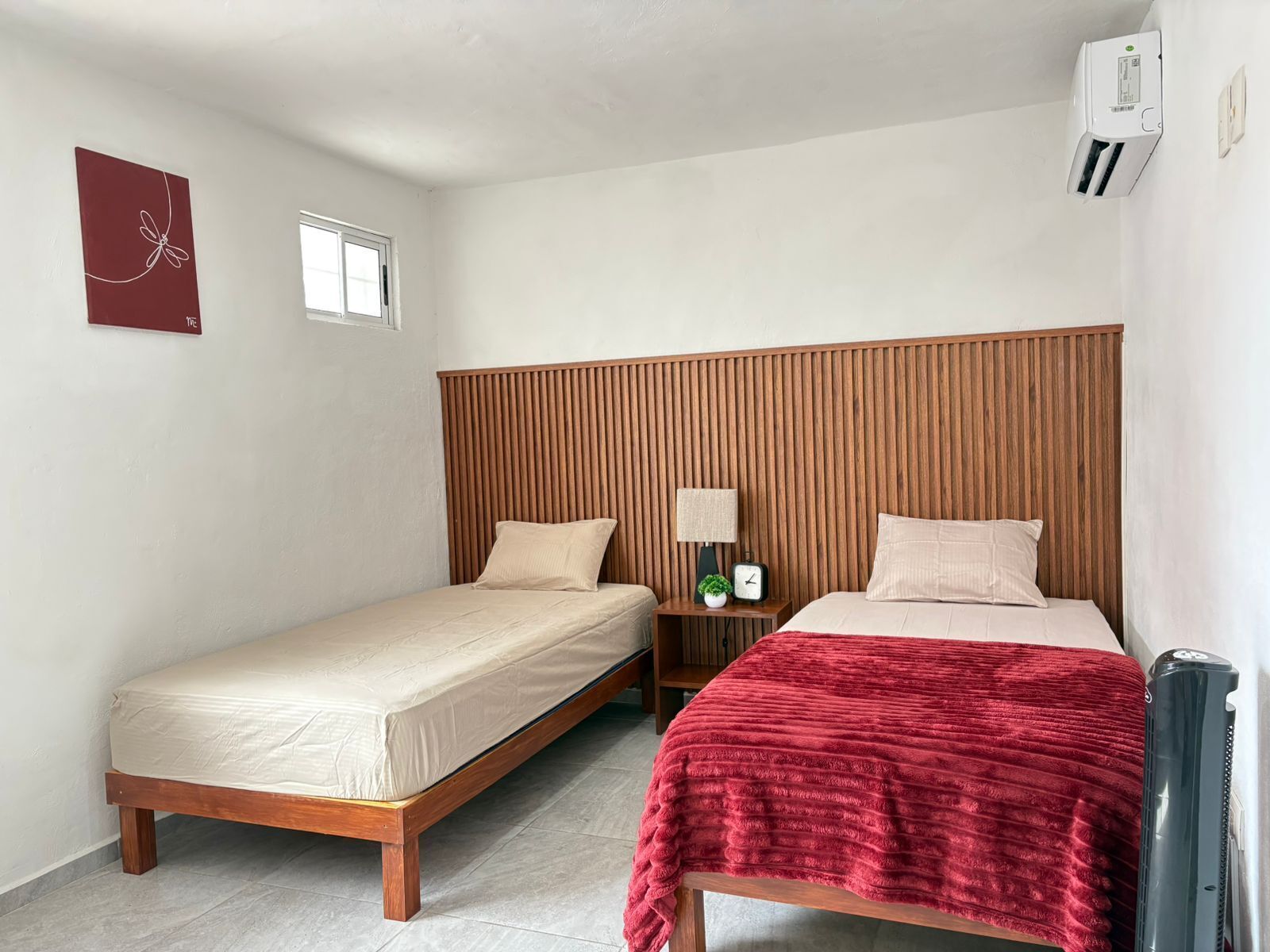 Una habitación con dos camas, un cabecero de madera, una mesita de noche con lámpara y un ventanal pequeño.