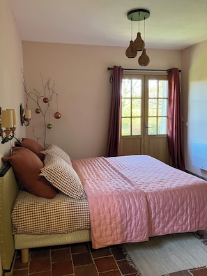 Una camera da letto con un letto, cuscini, una lampada a sospensione, una finestra con tende e una mensola a parete con oggetti decorativi.