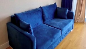 Um sofá azul num quarto com piso de madeira, um aparelho de ar condicionado na parede e uma janela com cortinas escuras.