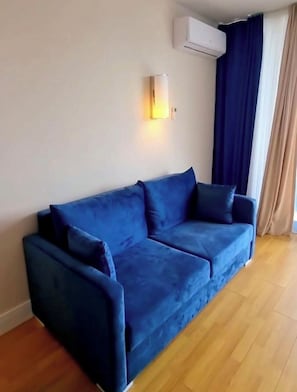 A blue sofa in a room with a wooden floor, an air conditioning unit on the wall, and a window with dark curtains. - Fantastic 1-bedroom apartment with AC Panoramic View (Batumi)