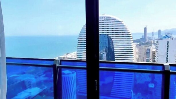 Vista de um edifício alto com varanda de vidro, com vista para a paisagem urbana e o mar.