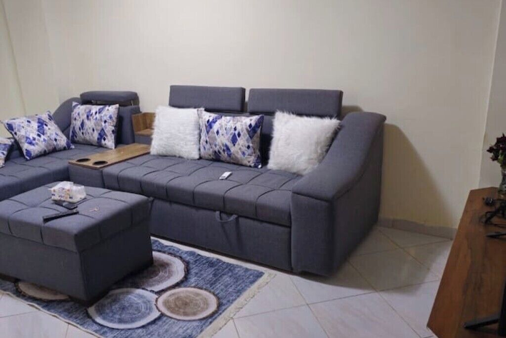 Un salon moderne avec un canapé d’angle gris, un pouf assorti et des coussins décoratifs à motifs. Une table basse en bois est placée devant le canapé, et un tapis bleu et blanc recouvre le sol.