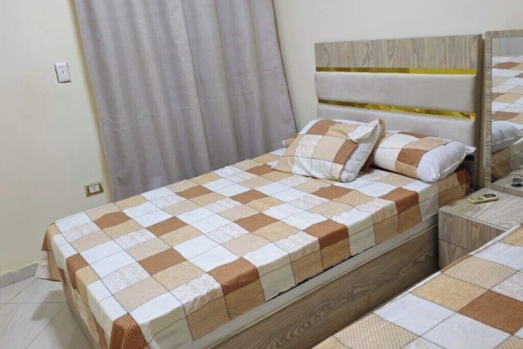 Un lit bien fait, recouvert d’une couvre-lit à carreaux, avec une tête de lit en bois et une table de chevet surmontée d’une lampe.