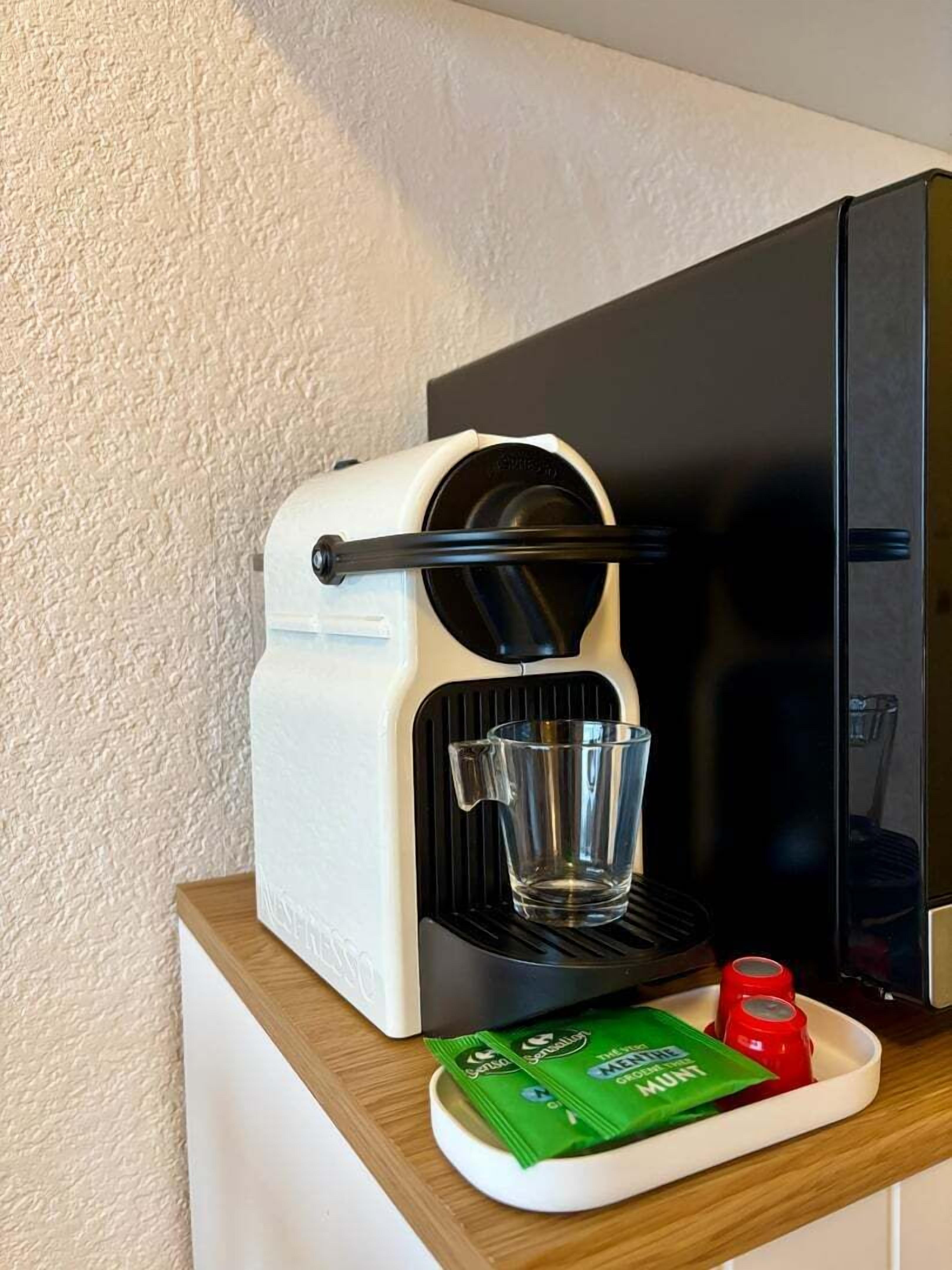 Een koffieapparaat met een glazen kophouder, een bakje met koffiecups en een rode fles.