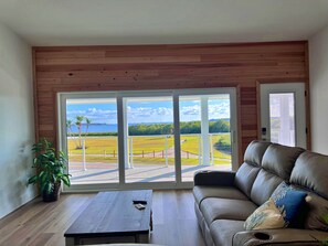 A living room with a leather sofa, a coffee table, and a view through large glass doors to a grassy area with a palm tree and a body of water. - Bay view home w/private hot tub, 2 real desks & fenced yard. 31+ night stays (Oldsmar)