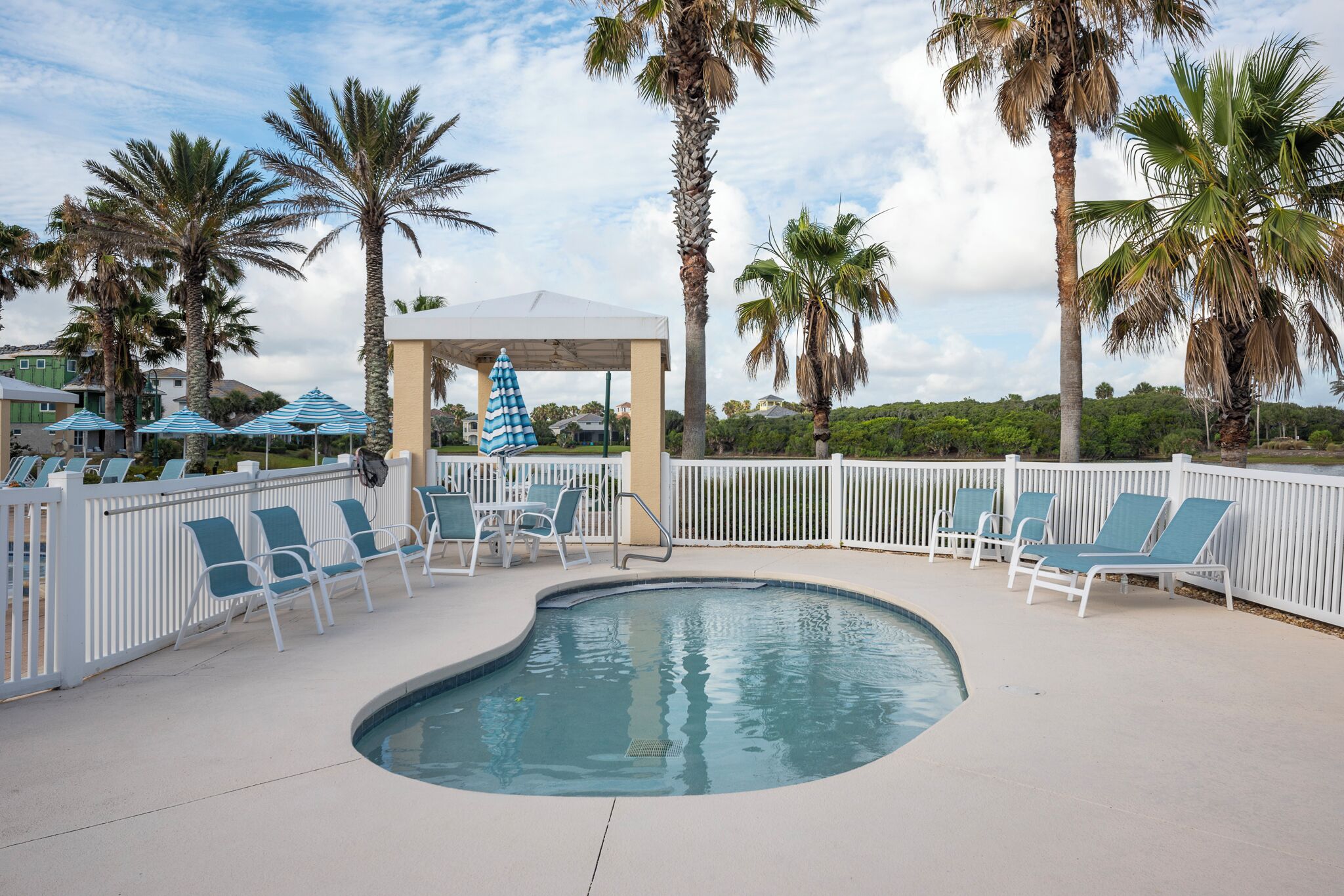 A pool area with lounge chairs, a covered seating area, and palm trees.